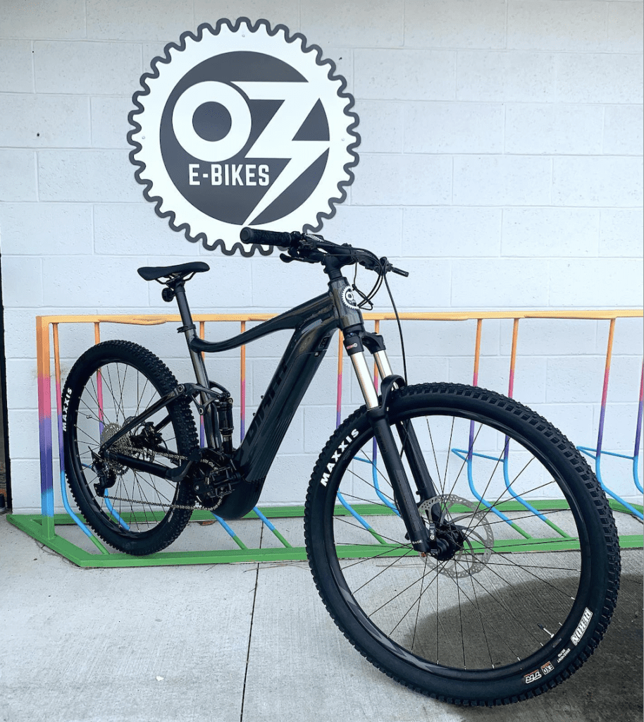 An ebike stands in a bike rack with an OZ E-bikes sign hanging on the wall behind it.