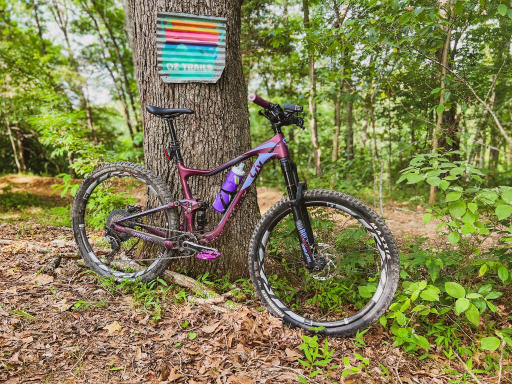 A purple mountain bike leans against a tree trunk with an OZ Trails signed pinned on it.
