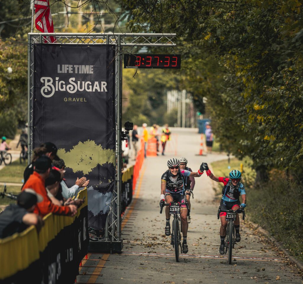Two female racers ride across the finish line holding their hands up together.