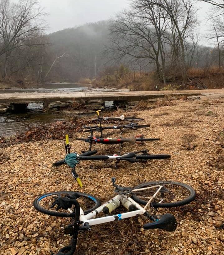 Five gravel bikes lay on the ground with a creek crossing in the background.