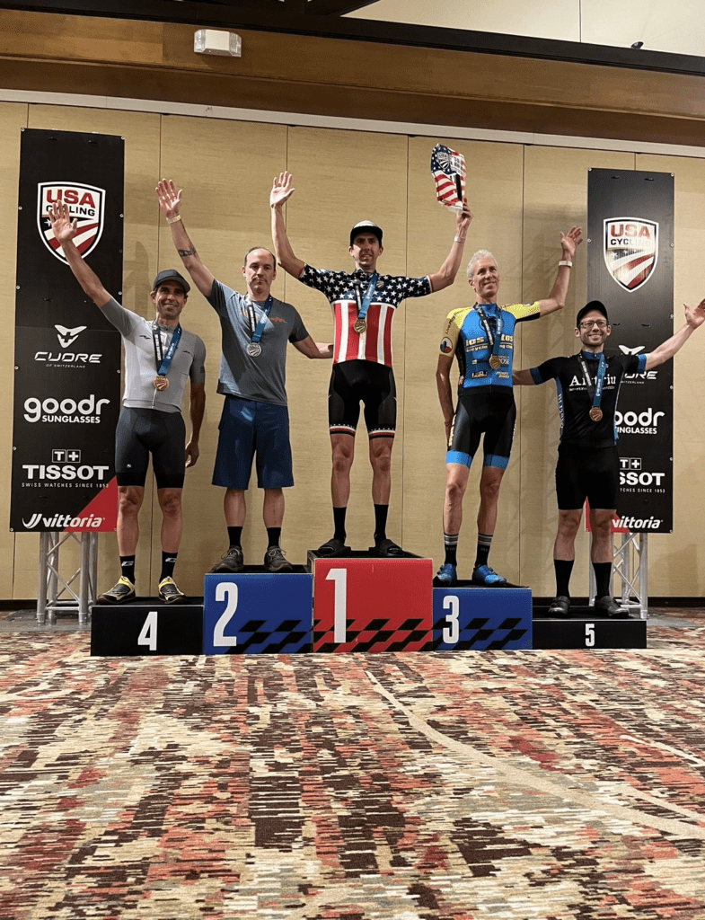 Bike School Bentonville Coach Chris stands on the top step of the podium. Places 1-5 are on the podium and Chris is in the middle on the top step.