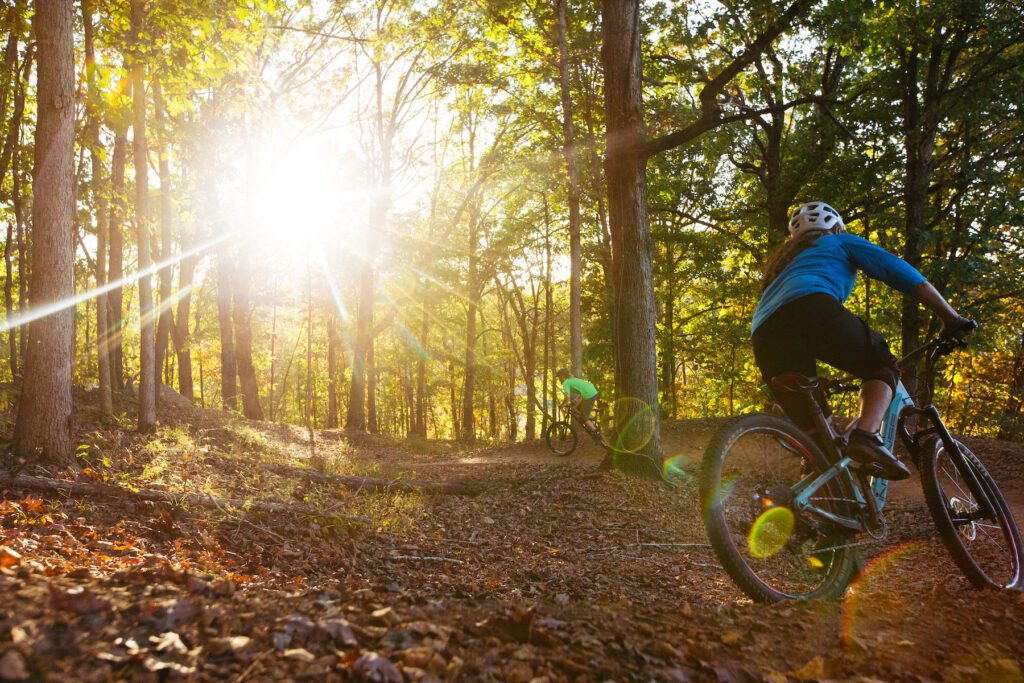 Two people ride a mountain bike trail with a bright yellow sunset in the bakcground
