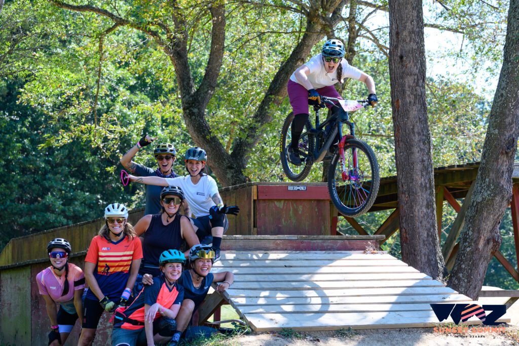 A group of female mountain bikers stand in a group and smile while another rider goes off a drop.